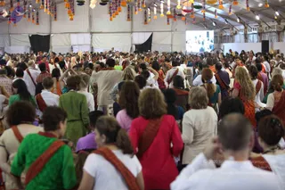 A large crowd fills a white tented hall hung with colourful paper lanterns and tassels overhead. Many devotees, most in saffron, orange, green, red and white clothing with saffron shawls over their shoulders, face away from the camera toward a distant figure near a golden ceremonial object at the far end. A large screen on the back wall shows a distant image. In the foreground, a woman in green and a woman in red stand among dozens of other participants. No individual person can be identified from their appearance.