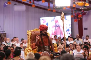 A seated figure on an ornate gold-and-brown throne at the front of a hall, wearing a red-orange turban with a jewelled ornament, orange-saffron robe and leopard-print lower cloth, with long rudraksha-style beaded malas and a rudraksha-looking necklace. He gestures with one raised hand while sitting beside a carved golden lion figure. A tall silver trident stands to his right. Behind him a large screen shows a figure in red robes, and the audience in the hall wears white, with orange and yellow tassel decorations hanging above. Swamiji (probable): the seated figure has the saffron robes, red turban and long malas of a candidate. A crowd of dozens of people is visible.