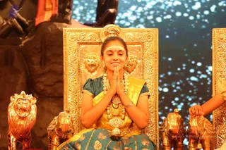 A young woman sits on an ornate gold throne with carved lion heads and gold lion and elephant figurines at its base, pressing her palms together in anjali mudra with her eyes closed and a smile. She wears a teal and gold silk sari, gold necklaces and a layered gold pendant, bangles, and a white tilak on her forehead with her hair pulled back. Behind her a large dark sculpted figure and a blue starry backdrop fill the stage. Another person's hand rests on the adjacent throne at the right edge.