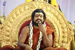A man sits on an ornate gold throne with a red cushion, smiling with his hand resting on his chin. He wears an orange-saffron cloth over his shoulders and a long garland of white flowers with blue and gold beads. A white and red forehead marking is visible, and his bare arms show stripes of white ash. Behind him are gold decorative elements and a pale lavender backdrop. Swamiji (probable): a bearded figure with long swept-back hair, saffron wrap and ceremonial garland seated on a throne. The image carries a faint copyright line along the lower right.