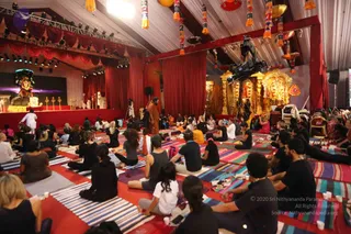 A photograph of a large tented hall with red drapes and hanging colourful tassel decorations. Many participants sit cross-legged on striped rugs and colourful mats, mostly in black and grey clothing, facing a central figure in an orange-brown robe standing near the middle of the floor. To the right stands an ornate gold shrine with a deity murti, and a film camera on a crane is mounted overhead. On the left a stage holds a decorated altar with a deity image and brass lamps, and a white-clad figure stands near it. Participants appear to be in a group yoga or meditation session.