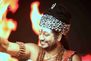 A smiling man with long dreadlocks wrapped in a leopard-print headband with a small trident-like ornament at the top. He has a red tilak on his forehead and wears multiple strands of rudraksha beads, a gold chain, and a red-orange garment over his shoulder. His right arm is extended toward the left edge of the frame, with rudraksha bracelets on the wrist. Behind him, large orange and yellow flames rise against a dark background.