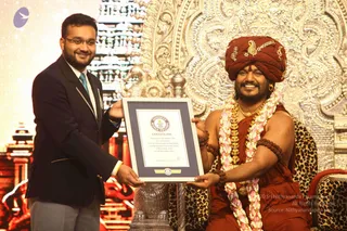 A man in a dark navy suit, light blue tie and grey trousers stands at left, smiling, holding a framed Guinness World Records certificate. Beside him, a seated man in a red-orange turban with gold ornaments wears an orange-red robe, multiple long pink-and-white flower garlands, a gold necklace and rudraksha bracelets, and smiles broadly. He sits on a leopard-print cushioned seat in front of an ornate gold carved backdrop with a silver-toned decorative stand below. Appearance-based candidate: Swamiji (probable), seated in the honoured position, with a crown-like turban, ornate gold adornments, orange robe and garlands, consistent with the page. The certificate reads Guinness World Records and certifies a record achieved by H.H. Sri Nithyananda Swamiji and Nithyananda University at Bengaluru.