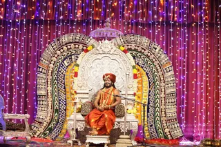 A seated figure sits on an ornate silver throne with a large jeweled arch behind it, decorated with colorful beaded and sequined panels in green, red, yellow and blue. Above the throne hangs a silver ceremonial canopy, and a microphone stands in front. The seated person wears an orange-red turban, an orange-saffron dhoti-style garment, a bare upper body with bangles, and a long yellow-and-orange flower garland with a rudraksha-style mala draped around the neck. Strings of multicolored fairy lights and red-purple drapes frame the stage, and brass oil lamps sit on the lower right.