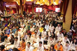 Large indoor satsang hall decorated with yellow and red drapery and hanging yellow lanterns. Hundreds of devotees sit cross-legged on orange and red mats, many wearing white kurtas or white clothing with rudraksha malas. Some wear orange or saffron garments and others colourful saris and salwar kameez. Two large screens on the back wall show a figure in red and white, and a deity murti stands on a decorated altar at the left. Watermarked copyright text appears in the lower right corner.