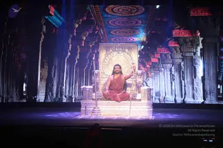 A seated man in red-orange ochre robes sits cross-legged on an ornate golden throne on a stage, holding a tall golden staff in his right hand. He wears a red-orange shawl, a rudraksha mala and a gold necklace, with long swept-back hair. Behind him a large projected backdrop shows a temple hall with carved pillars, lotus-like rosettes and a richly patterned ceiling in purple, blue and red, lit by blue and purple stage lights. a person is seated on the throne, identified by his distinctive hair, saffron robes, rudraksha mala and gold necklace. The image is a stage performance scene in a temple-style set.