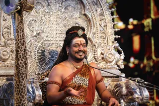 A man seated on an ornate silver throne with embossed floral and animal-motif carvings speaks into a microphone while smiling and gesturing with his right hand. He has long matted hair with a red and gold headpiece, white and red tilak on his forehead, a gold necklace with a pendant, and wears a leopard-print cloth over an orange-red shawl, with red rudraksha bands on his arms and wrists. A leopard-print cloth hangs over the left side of the throne and a blurred red-and-gold backdrop lights the right. Only one person is visible.