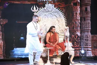 A stage event with an ornate silver throne carved with floral and deity motifs, set against a backdrop of red temple-style carved pillars. A seated figure on the throne wears a saffron-orange robe and multiple rudraksha malas, with a white tilak on the forehead and long, swept-back hair, and has a crown-like ornament on the throne above. A standing man in white kurta and white trousers, wearing a rudraksha mala, holds a small cup and smiles toward the seated figure. A silver trident stands behind the throne, and a microphone stand is at right. Chandeliers hang overhead, and a blurred head of another person is visible in the lower foreground. Swamiji (probable): the seated figure shows long swept-back hair, saffron robes and multiple rudraksha malas on a throne.