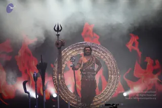 A bare-chested man stands within a large ornate golden ring on a stage filled with smoke and red-orange flame backdrops. He wears a tiger-print or leopard-patterned waist wrap, a dark rudraksha-style mala and ceremonial beaded adornments across his chest, with a red tilak on his forehead and long swept-back hair. He holds a long staff-like mace in one hand and rests the other on his hip. To the left stand ceremonial weapons: a tall trident, a dark ceremonial sword or kukri, a curved blade and a feathered ritual staff. Stage lights and a camera rig are visible at the bottom. The setting resembles a dramatic theatrical or devotional performance. a person: the figure's long hair, bare upper body with adornments and ceremonial staff correspond to the event's presented figure, although appearance alone is limited.