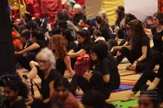 A photograph of a large seated meditation or yoga gathering on colourful mats in a hall. Many participants wear black tops and black trousers and sit cross-legged or with legs folded to the side, eyes closed or looking down. In the foreground a woman with silver-white hair and glasses sits beside a woman with long reddish-brown curly hair. A red backpack rests among the mats. At the upper left, figures in red and orange robes sit near a gold-coloured seat, and a small bottle of water stands on the floor at lower right. Some participants are partially visible at the edges of the frame.