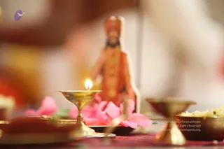 A brass oil lamp with a lit flame stands in the foreground on a red cloth, beside pink flower petals and a brass bowl of white rice or offering. Behind them, a small blurred figure in saffron-orange robes with a long beard and a tall crown-like headdress sits as a depiction. The background is softly out of focus with warm tones. The setting suggests a puja arrangement with lamp, flowers and offerings.