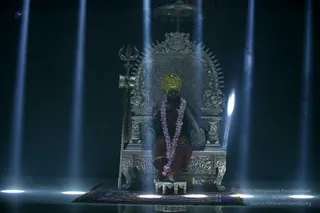 A seated figure on an ornate silver throne (peetha) in a dark hall, lit by vertical blue beams of light. The figure wears a golden crown, a garland of purple and white flowers, and a rust-red-brown robe, with a trident-shaped staff and a silver ceremonial umbrella beside the throne. The figure's feet rest on a small silver footstool atop a purple and gold patterned rug. The scene resembles a staged presentation rather than a crowd event.