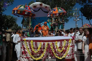 A street procession in daylight with a decorated vehicle at the centre. A figure in orange robes sits on a raised float under two ornate umbrellas with tasselled edges, one printed with a dove and the word Dhyanapeetha-style lettering. A woman in orange with a beaded necklace sits beside a bald man in orange robes, and a bare-chested man sits in front of the float. A tall silver trident stands at right. The float's white and red cloth is draped with yellow and pink marigold and rose garlands. Around the vehicle, many men in white shirts, orange shawls and khaki uniforms walk and crowd close, and a woman in white with an orange shawl stands at right. Trees and blue sky are behind.