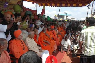 Outdoor gathering under a red and yellow striped canopy on a red carpet. Several saffron-robed sannyasis sit in the front row; the foremost on the left wears an orange turban and a rudraksha mala, and a central saffron-robed figure with a grey beard sits with hands folded. Beside them a man in cream clothes sits in a chair. Behind them, devotees hold green and terracotta kalasha pots raised overhead, and a man in a black shirt holds a microphone. A standing man in a green striped shirt faces the group. Yellow and red banners and a petrol station are visible in the background, with a crowd of onlookers beyond.
