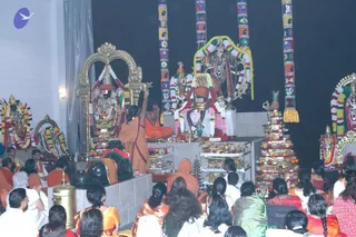 Photograph of a temple puja inside a hall with several large deity murtis decorated with flower garlands and colourful striped pillars above. In the centre, a standing priest in a saffron-orange robe raises a lit lamp toward a decorated altar, while another orange-clad figure sits cross-legged beside him. Tiered trays of fruit and sweets stand on the right. Many seated devotees in orange, saffron, red and green saris and white dhotis face the altar, some with hands raised. The scene resembles a puja or aarti. No candidate is clearly identifiable from appearance, so the a person field is unsure.