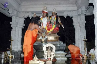 A photograph of a temple shrine at night showing a large stone-and-metal murti pair of deities, one with a crown and red headband and one with a crown and red-and-green sari, both garlanded with yellow and red flower garlands. In the foreground a shirtless man with long dark hair in an orange-saffron dhoti stands bending toward the base of the murti, pouring or applying offerings from a metal vessel, as an abhishekam-style puja is performed. A second person in orange clothing kneels at the right on the wet floor, and a figure in white stands at the far right edge. Brass oil lamps flank the base and metal pots sit at the left. A smaller brass deity figure sits on a small altar in the lower centre. Grey carved pillars frame the scene.