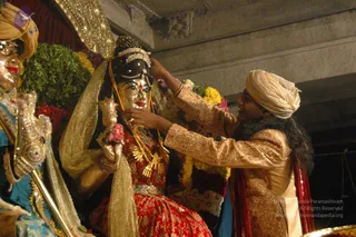 A photograph of a ceremonial wedding ritual at night, centred on a golden-faced deity murti of a goddess with a dark hair bun, gold crown, red-and-gold sari and a gold garland-like necklace, holding a pink flower. A man in a cream turban and a gold brocade sherwani-style tunic with red sash stands at right, adjusting the goddess's veil and tying or arranging her gold necklace with both hands. Behind them are garlands of yellow, orange and red flowers and green leaves, with an ornate gold-framed canopy above. A partly visible figure in a blue garment and turban with a gold ornament appears at left edge. The scene suggests a thirukalyanam-style marriage ceremony.