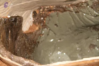 Close-up photograph of a wooden trough or vessel filled with a thick, pale grey-green paste, possibly a clay, mud or ritual mixture, with glistening droplets and flecks of reddish-brown material along its edges. No people are visible; the frame shows only the container and its contents against a pale, textured surface.