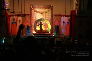 Photograph of an evening stage performance in a hall, viewed from the audience. A performer in dark costume with a red wrap stands in front of a large glowing green and yellow ring, with an orange flame-like light behind. Red and white draped curtains, a garland of marigolds across the back panel, and decorated side panels frame the stage. Silhouetted audience members, including a raised arm in the foreground, fill the bottom of the frame. A seated figure in pale clothing sits at the stage edge and two people in white stand near the stage. Ceiling fans hang above.