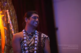A photograph of a smiling person standing at the side of a stage in front of a dark red curtain, with a gold-textured decorative panel on the left and a plain pale wall on the right. The person has long dark hair pulled back, a small mark on the forehead, and wears a sleeveless top with a zebra-stripe pattern in black and cream. Layered around the neck are multiple strands of rudraksha beads with white skull-shaped beads and a dark pendant. Warm purple and orange stage lighting falls across the scene. Swamiji (probable): long swept-back hair, rudraksha malas and ceremonial ornamentation identify this person as a.