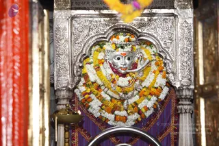 A silver-faced deity murti is framed in an ornate silver niche within a temple shrine. The murti is heavily garlanded with white and orange-yellow marigold garlands and wears silver ornaments and a silver crown; a red-orange tilak marks its forehead. Above it, an inscription is carved in stone, and a flower garland hangs at the top edge. Below, a brass oil lamp with a flame stands before a silver rail, and a purple cloth with gold trim drapes the base. Red and gold panels frame the sides. No people are visible.