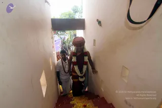 A photograph taken looking down a narrow whitewashed stairwell with red-painted steps strewn with yellow flower petals. A central figure in a maroon robe with an orange-red turban and a long garland of white and red flowers walks down the steps, one hand resting on the wall. Behind them a man in a white shirt and white dhoti, with a red shawl draped over his shoulder, follows, and another figure in orange clothing is visible further back. Light enters from an open doorway at the top with greenery visible outside.