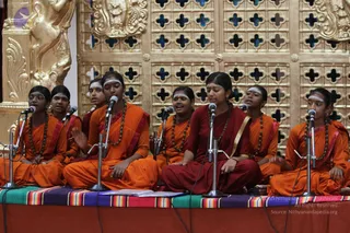 A group of young women in orange and maroon robes sit cross-legged on a stage with a multicoloured striped cloth, singing into standing microphones. Most wear rudraksha malas and white forehead markings, and one in a deep red robe with a gold sash sings at centre. Behind them is an ornate golden lattice screen with blue cutouts and gilded carved figures on the left. The group appears to be performing chanting or devotional singing.