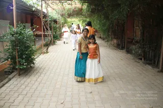 Photograph of a paved pathway shaded by overhanging bamboo and climbing vines, with a red-brick building on the left. In the foreground, a smiling young woman in a teal skirt and gold-bordered cream blouse stands beside a girl in a white skirt with gold border and an orange-gold top, both posing for the camera. Behind them, several people in white and orange clothing walk along the path, including a figure in white with a red sash and a figure in orange robes.