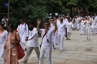 A large group of devotees walks along a stone-paved path beneath green trees, many dressed in white. A man in the centre wears a white polo shirt, white trousers and a red lanyard, raising both open hands in greeting while smiling. Nearby a woman in a peach sari with a red-orange bag, and another man in a white shirt with a red lanyard, walk among others in white kurtas, dhotis and lanyards. Some participants wear orange-red garland lanyards and name badges. The procession suggests an ashram gathering or walking procession.