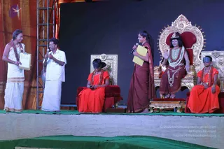 Stage event with a green-edged platform in front of a dark blue backdrop and a red-and-orange drape at left. On the right, an ornate gold throne with a red seat holds a figure in a saffron-orange robe, a crown-like headpiece, layered beaded necklaces and a rudraksha-style mala; the figure appears to be a statue or mannequin with a painted face. A black-blindfolded young person in orange-red robes sits on a smaller throne to the centre-left and another blindfolded child in orange-red sits at far right. A woman in a dark red sari holds a microphone and a yellow folder, while a man in white dhoti and shirt holds a microphone beside a man in white clothes with a clipboard at far left.