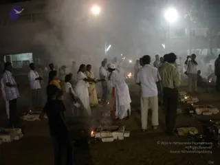 Night outdoor fire ritual in a sandy compound lit by floodlights, with smoke drifting through the scene. Several small brick-lined fire pits glow with flames and oil lamps on the ground. People stand around them: a woman in a white sari holds an offering near a fire, a figure in white with an orange sash stands at the centre, and men in white and light shirts and trousers watch. A man at right in a dark jacket and a person with a camera stand nearby. Pale brick structures and a building with windows form the background.