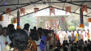 A photograph of a crowded outdoor temple-festival scene under a corrugated roof strung with orange and saffron pennants and tall striped pillars. A large deity murti or decorated image sits in a shrine framed by palm fronds, red cloth and heavy garlands, with a priest in a white dhoti and orange shawl standing nearby. In the foreground, devotees crowd forward, including a woman in a blue top with a long braid, women in saris, and men in white dhotis and shirts, some wearing flower garlands. The scene appears to be a procession or darshan gathering during a temple festival.