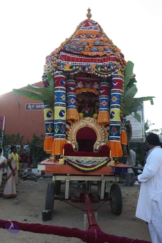 A richly decorated festival chariot (ratha) with a tiered, multicoloured canopy of red, orange, yellow and blue cloth with floral and bird motifs, topped by a golden finial and hung with marigold garlands. Banana leaves frame the sides. Inside the open shrine stands a deity murti with a Shiva lingam and bull motifs on the back panel, and an empty ornate gold-and-red throne sits at the front of the platform decorated with flower garlands. A woman in a cream sari with a yellow shawl stands at left, and a man in white kurta stands at right near the chariot. A red rope barrier runs along the foreground. A red building with a Kannada sign and a banner reading Nithyanandapuram-style signage appears behind.