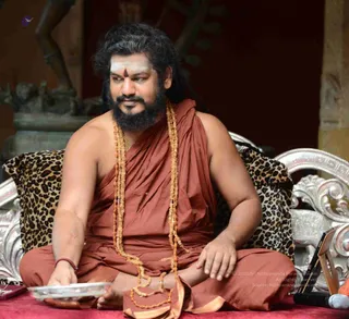 A photograph of a bearded man with long dark hair seated cross-legged on a silver-framed chair with a leopard-print cushion, set on a red cloth. He wears a rust-brown shawl draped over the shoulder and a long layered rudraksha mala with ornamental gold necklaces, with white vibhuti and a red tilak on his forehead. He rests one hand on a silver tray, and a microphone and small equipment sit at the lower right. Behind him a bronze sculpture stands on a dark background.