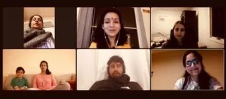 A six-panel video call grid of participants at home. Top left, an older woman with grey hair wrapped in a grey blanket. Top centre, a woman in a yellow top with earphones and a red bindi smiles at the camera. Top right, a woman sits on a bed in a plain room. Bottom left, a young boy in a green shirt and a woman in a pink top sit together on a sofa. Bottom centre, a man in a dark north-face hoodie with headphones sits in front of a white curtain. Bottom right, a smiling woman in glasses and a white patterned top. No a person candidate is visible; the screenshot shows only a remote gathering of participants.