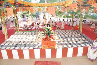 An outdoor ritual space under a canopy hung with orange and yellow pennants and banana-leaf decorations, with a red carpet on the ground. In the foreground, a large deity murti dressed in green and red garments with a white floral garland stands on a raised platform, surrounded by rows of metal plates heaped with food offerings and small bowls. Behind the platform, a few dozen people in white dhotis and white saris sit on the carpet or stand, some with their hands folded. A red and white striped cloth covers the platform's base. On the far right edge, a woman in a white sari with a purple border stands near the table.