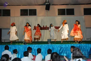 A photograph of a children's performance on a stage with a blue draped skirt and green mat. Six young women in white and orange dance in a line before a white curtain, with red letters behind them reading partially "Ananda..." and "2010". Several dancers wear orange or red headscarves and sheer orange shawls over white clothing. Seated young children in white school uniforms watch from the front edge of the stage. No a person candidate is visible on the stage or among the audience.
