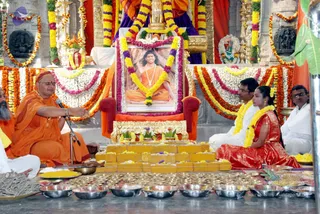 A photograph of a ceremonial puja in an ornately decorated hall with marigold and jasmine garlands draped along the walls and altar. At the centre, a large framed portrait of a seated man in saffron robes and a white bead necklace is garlanded with yellow and purple flowers and set on a red and gold throne. In front of it stands a tiered arrangement of yellow-cloth-covered steps with brass plates and pots of flowers. A saffron-robed man with a shaved head and a red headband sits cross-legged at a microphone, gesturing with his right hand and holding a tray of yellow powder. Two people sit to the right: a man in white clothes with a yellow-and-white garland, and a woman in a red silk sari with a yellow garland, seated in front of rows of small oil lamps and metal bowls. A bearded man in saffron is partly visible at the far left, and another man in white stands at the far right.