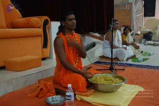 A photograph of a satsang hall where a man in orange robes and a rudraksha mala sits cross-legged on an orange mat, gesturing with his hands at a microphone on a stand. He has long dark hair and a red tilak, and wears a gold necklace and orange wrap. Beside him a bald man in white dhoti and shawl with a gold chain and tilak sits, and a woman in a yellow sari sits nearby. A large steel bowl of yellow rice, small steel bowls, a water bottle and banana-leaf plates lie on the mat. Behind them stands an orange cushioned throne and a cream chair. Swamiji (probable) is the man in orange with the long hair, mala and gold necklace, seated centrally as the speaker.