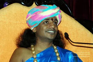 A photograph of a man seated before an ornate golden-upholstered chair with dark carved wooden arms and a red velvet curtain behind. He wears a multicoloured pink, turquoise, yellow and white turban, a red vermilion dot on his forehead, a blue silk shawl and a gold bead necklace with large round beads. Long dark hair falls past his shoulders and a microphone on a stand is visible at right. He is smiling and looking up to the side.