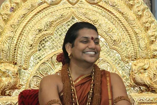 A smiling man with long dark hair swept back sits in front of an ornate gold throne with carved floral and bird motifs. He wears a saffron-orange shawl over his shoulder, layered rudraksha mala necklaces, a small gold stud earring, a red bindi on his forehead, and rudraksha bracelets on his upper arms. A red cloth with a tiger-stripe pattern rests beside him. The long hair, ornate gold seat and saffron clothing with rudraksha malas give appearance-based cues for Swamiji (probable), seated in person during an event.