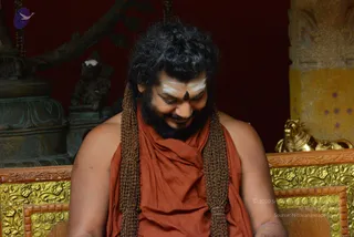 A man with long, dark, curly hair and a full beard sits on an ornate golden throne, looking down with a gentle smile. He is bare-shouldered, wearing a rust-orange cloth draped over his body and two long brown rudraksha-style mala strands hanging down his chest. A white tilak and a dark red dot mark his forehead. Behind him stand a bronze figure and a golden ornamental pillar against a deep red wall. The image is a photograph; the appearance matches the a person candidate but identity is uncertain, so the role is described as a seated devotee and teacher.