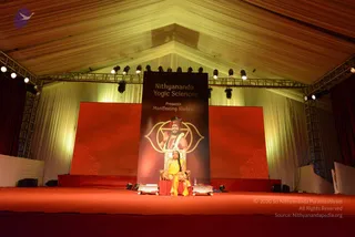 A stage under a tent at an indoor-style event, with red carpeting and a red backdrop. A tall vertical banner reads Nithyananda Yogic Sciences Presents Manifesting Shaktis and shows a figure in a red turban and white garland beside a chakra-style emblem. Below it, a woman in a yellow sari with a long braid sits on a red-cushioned seat between two small decorative stands, with a water bottle nearby. Stage lights hang from the metal truss overhead. The stage is otherwise empty and no audience is visible.