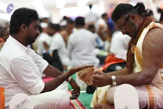 A seated man in a white shirt and white dhoti sits on a green-and-pink striped mat, extending his right hand toward a brown coconut held by a second man. The second man wears a wrap with yellow-gold borders over bare shoulders, a long rudraksha mala, a round gold-toned pendant, a silver bracelet, and eyeglasses, with white ash marks on his chest and arms. He bends over the coconut in a focused gesture, hands clasped around it. A small white paper plate with the handwritten name Priyan lies in the foreground, and a blurred crowd of white-clad participants sits in the background of a large hall.