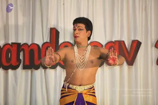 A performer stands center stage in a bare-chested costume, painted with dark eye makeup, red lips and a small tilak on the forehead. He wears multiple strands of white pearl necklaces, a green beaded collar, gold armbands and bangles, and a gold belt over striped orange and purple lower garment. His palms are raised outward in a dance gesture, one hand decorated with red henna. Behind him hangs a white drape with large red and dark lettering reading part of a word that is cut off at the frame edges.