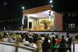 Night photograph of an open-air evening satsang on a lit stage in front of a large seated crowd. The stage has a banner reading in Tamil and English 'Nithyananda Dhyanapeetam, Tiruvannamalai' with red, gold and cream trim. Behind it a central throne-like seat is decorated with green and orange floral garlands, and a figure in orange robes sits on a golden chair at the centre of the stage. Strings of paper decorations hang above, bright floodlights and speakers stand at the sides, and the audience in white, pink, green and orange clothing sits on rows of chairs below.