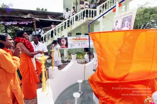 Outdoor photograph at a white building with a staircase and balcony, where a crowd of men in white shirts watches from above. In the foreground, a figure in orange robes with a red-and-white flower garland stands beside a brass oil lamp with a lit flame, while an orange cloth is draped over a white stone structure with a round dark basin. Two men in orange robes stand at left with hands pressed together in greeting. A cameraman in white clothing films the scene. A banner with Tamil text and a photograph hangs on the building, and two framed portraits are mounted at upper right. A blue-and-white pole and hanging orange cloth appear at the top.