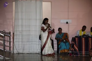 A woman in a white saree with a maroon-red border stands at left holding a microphone in one hand and a book in the other, speaking to an audience. Beside her, a seated older woman in a teal-blue saree with a gold border listens quietly with her hands in her lap. At right, two men in white with yellow shawls sit behind a table draped in a striped cloth, with microphones and audio equipment on it. A white curtain hangs behind the speaker against a pink wall, with metal railings at the left edge. The floor is polished tile.