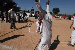 An outdoor photo of a tug-of-war contest on a sandy ground at a rural compound. A bald man in a white kurta with raised arms stands in the foreground, while another man in white trousers and an orange sash leans into a thick yellow rope. Several spectators in white and striped shirts stand near drum players with white drums, and a white van and a red flag are behind them. Some people in white sit in a row on a low wall in the background, and a woman in orange stands nearby. Bright sunlight and long shadows show it is daytime. No identifiable a person depiction is clearly visible; the bald man in white is a participant.