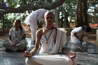 Outdoors under a large banyan tree, a young man with a shaved head sits cross-legged in meditation on a dark polished stone platform. He wears a white dhoti and shawl with an orange-and-green border, a rudraksha mala and a pendant, and has a vertical vibhuti or kumkum mark on his forehead with hands resting open on his knees. Beside him a woman in a grey-green patterned salwar and white scarf sits with hands folded in her lap, eyes closed. Behind them, a man in white bends forward in a bowing posture, and another person in white kneels in prostration at the right edge.